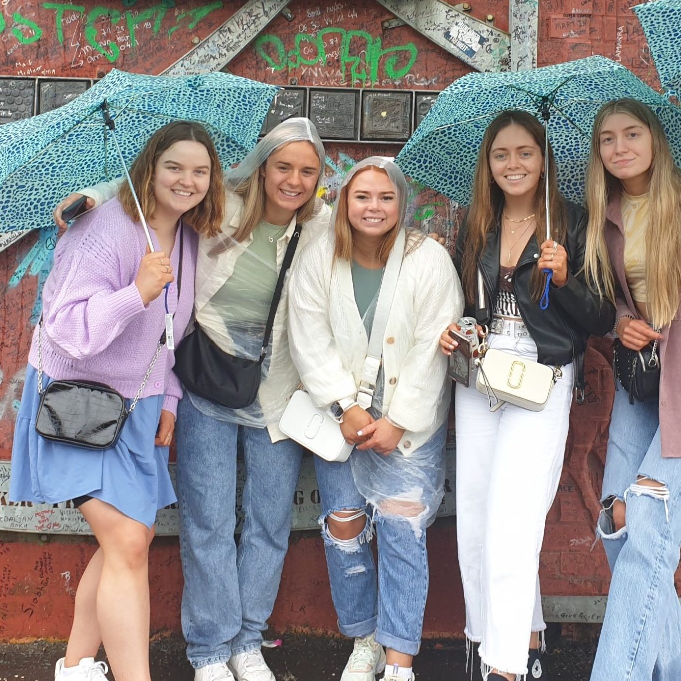 Peace Wall Belfast Group of five friends smiling, holding blue umbrellas in front of a colourful wall.